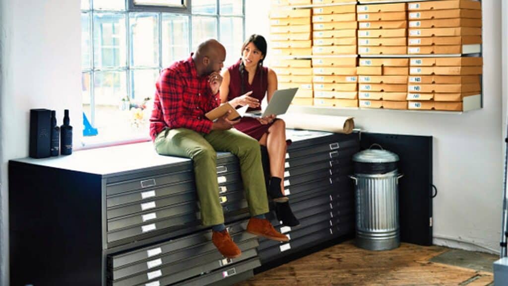 strategic compliance education with Man and woman sitting on filing cabinet and using laptop in modern office